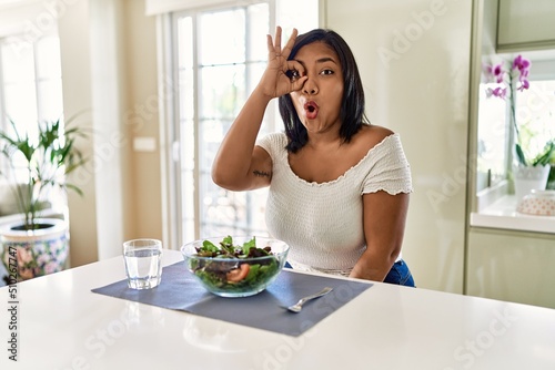 Young hispanic woman eating healthy salad at home doing ok gesture shocked with surprised face  eye looking through fingers. unbelieving expression.