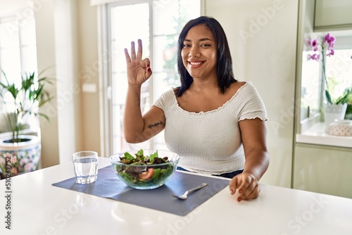 Young hispanic woman eating healthy salad at home smiling positive doing ok sign with hand and fingers. successful expression.