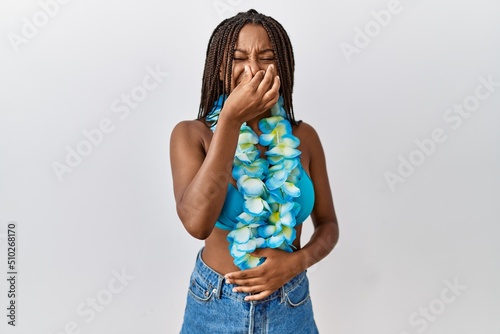 Young african american woman with braids wearing bikini and hawaiian lei smelling something stinky and disgusting, intolerable smell, holding breath with fingers on nose. bad smell