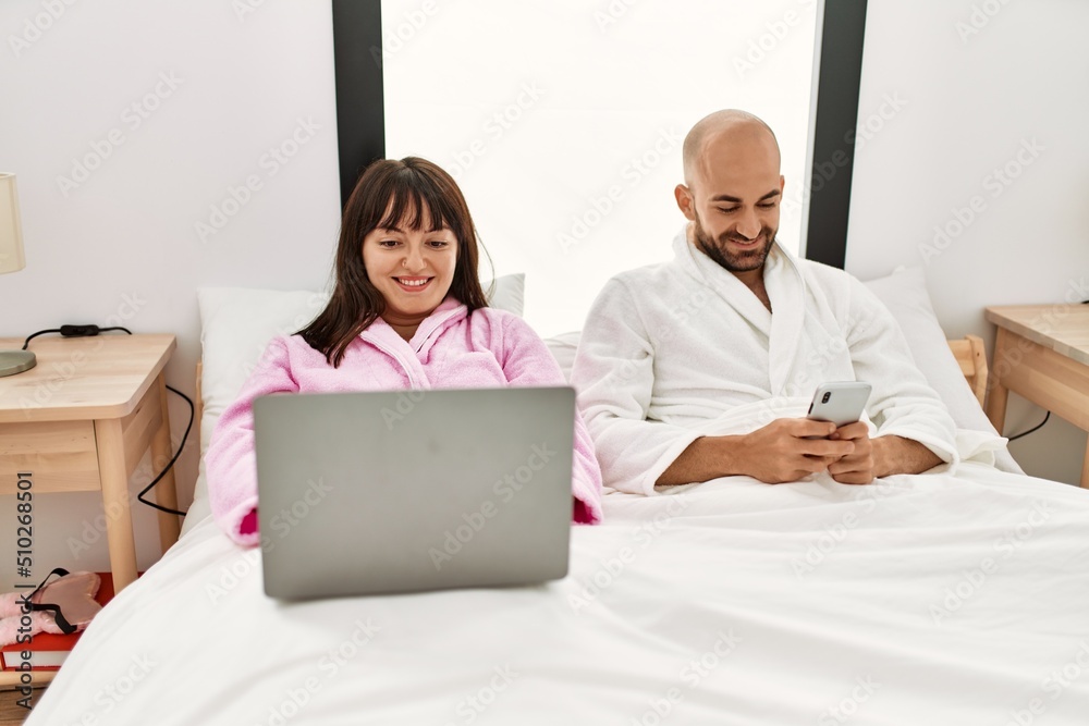 Obraz premium Young hispanic couple using laptop and smartphone sitting on the bed at bedroom.