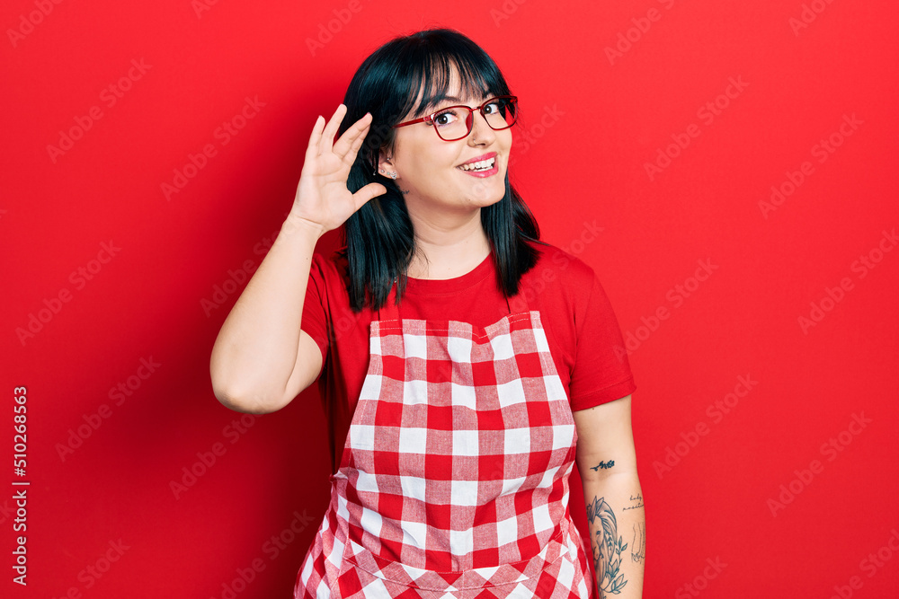 Young hispanic woman wearing cook apron and glasses smiling with hand over ear listening an hearing to rumor or gossip. deafness concept.