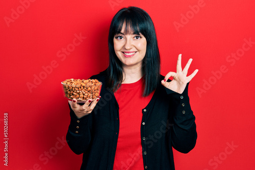 Young hispanic woman holding peanuts doing ok sign with fingers  smiling friendly gesturing excellent symbol