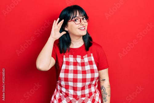 Young hispanic woman wearing cook apron and glasses smiling with hand over ear listening an hearing to rumor or gossip. deafness concept.