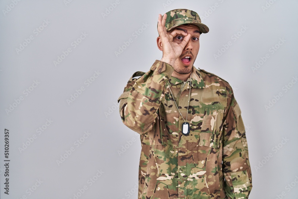 Young hispanic man wearing camouflage army uniform doing ok gesture shocked with surprised face, eye looking through fingers. unbelieving expression. Young hispanic man wearing camouflage army uniform doing ok gesture shocked with surprised face, eye looking through fingers. unbelieving expression.