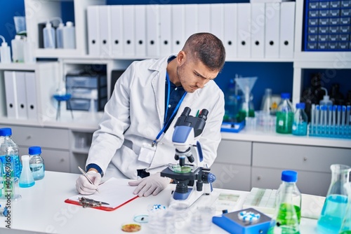 Young hispanic man scientist writing on document at laboratory