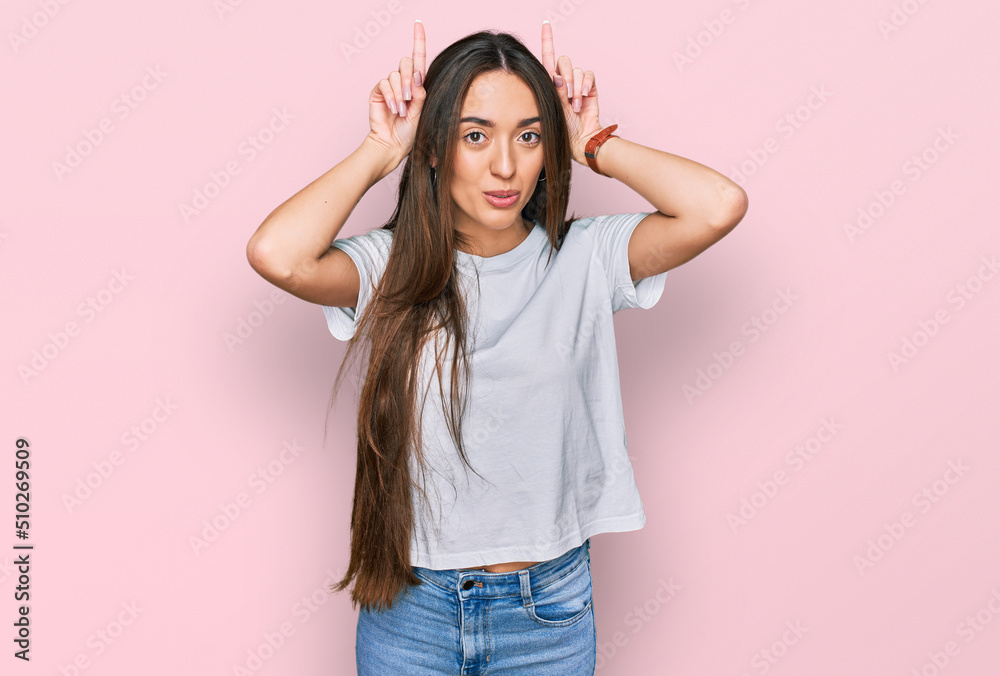 Young hispanic girl wearing casual white t shirt doing funny gesture with finger over head as bull horns Young hispanic girl wearing casual white t shirt doing funny gesture with finger over head as bull horns
