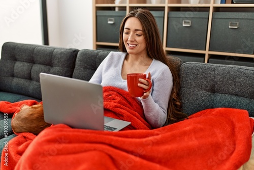 Photography Young hispanic woman using laptop and drinking coffee sitting on sofa with dogs