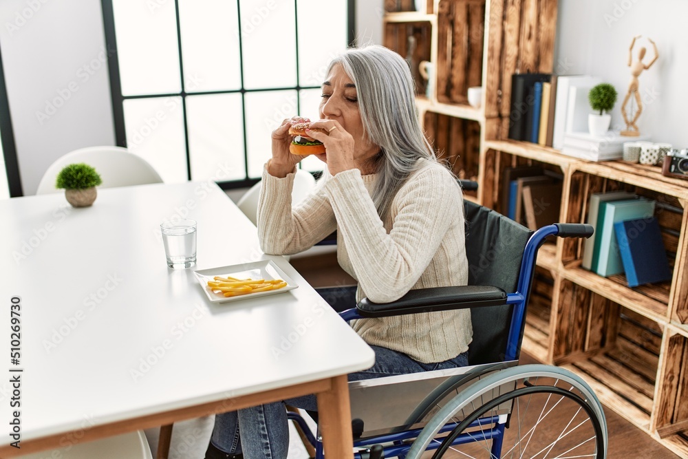Middle age grey-haired woman eating classical hamburger sitting on wheelchair at home Middle age grey-haired woman eating classical hamburger sitting on wheelchair at home