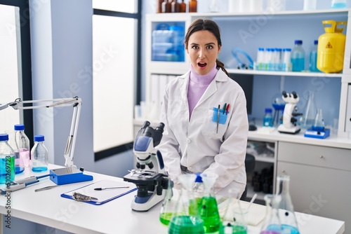 Young brunette woman working at scientist laboratory scared and amazed with open mouth for surprise  disbelief face