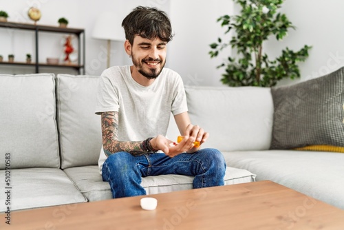 Young hispanic man holding pills sitting on the sofa at home