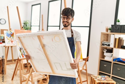 Young hispanic artist man smiling happy holding canvas at art studio.