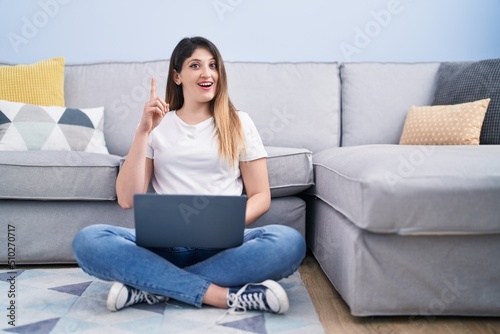 Young brunette woman sitting on the floor at home using laptop surprised with an idea or question pointing finger with happy face, number one