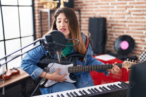 Young hispanic woman musician singing song playing electrical guitar at music studio