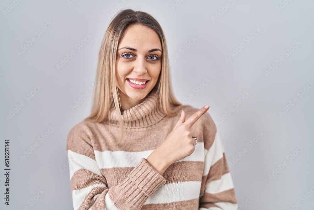 Young blonde woman wearing turtleneck sweater over isolated background cheerful with a smile on face pointing with hand and finger up to the side with happy and natural expression Young blonde woman wearing turtleneck sweater over isolated background cheerful with a smile on face pointing with hand and finger up to the side with happy and natural expression