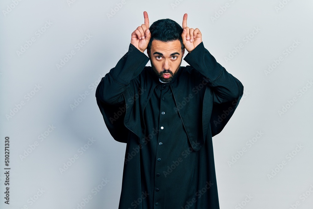 Handsome hispanic man with beard wearing catholic priest robe doing funny gesture with finger over head as bull horns Handsome hispanic man with beard wearing catholic priest robe doing funny gesture with finger over head as bull horns