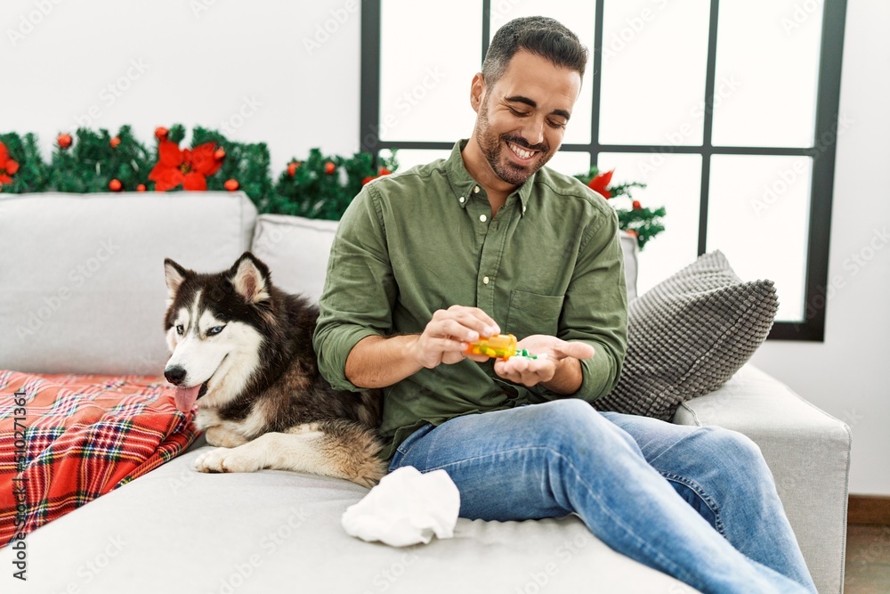 Young hispanic man taking pills sitting on sofa with dog by christmas decor at home