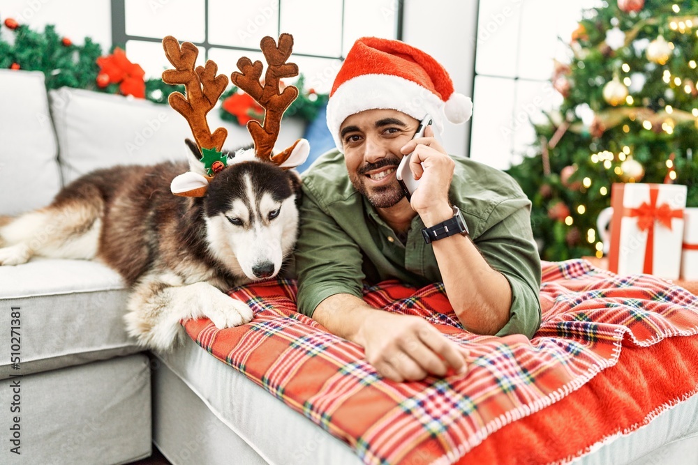 Obraz premium Young hispanic man talking on the smartphone lying on sofa with dog by christmas tree at home