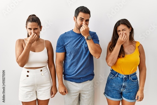 Group of young hispanic people standing over isolated background smelling something stinky and disgusting, intolerable smell, holding breath with fingers on nose. bad smell