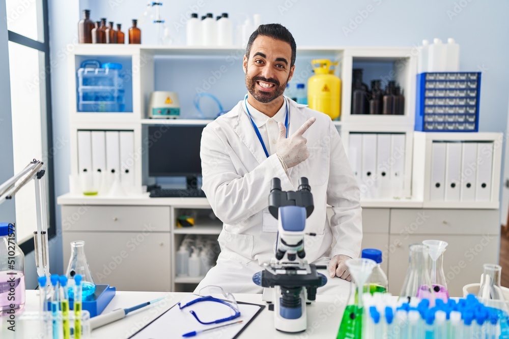 Young hispanic man with beard working at scientist laboratory cheerful with a smile of face pointing with hand and finger up to the side with happy and natural expression on face Young hispanic man with beard working at scientist laboratory cheerful with a smile of face pointing with hand and finger up to the side with happy and natural expression on face