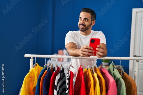 Young hispanic man using smartphone leaning on clothes rack at laundry room