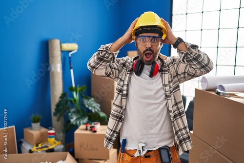 Young hispanic man with beard working at home renovation crazy and scared with hands on head, afraid and surprised of shock with open mouth