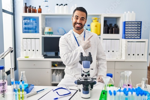 Young hispanic man with beard working at scientist laboratory cheerful with a smile of face pointing with hand and finger up to the side with happy and natural expression on face