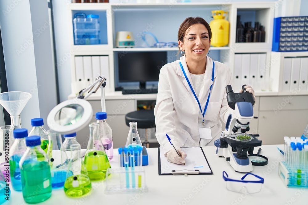 Young hispanic woman wearing scientist uniform using microscope and writing on clipboard at laboratory