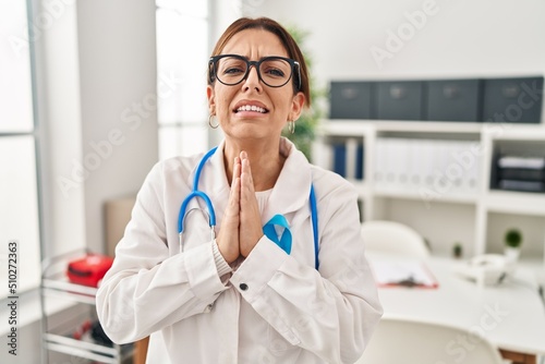 Young brunette doctor woman wearing stethoscope at the clinic begging and praying with hands together with hope expression on face very emotional and worried. begging.