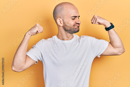 Young bald man wearing casual white t shirt showing arms muscles smiling proud. fitness concept.