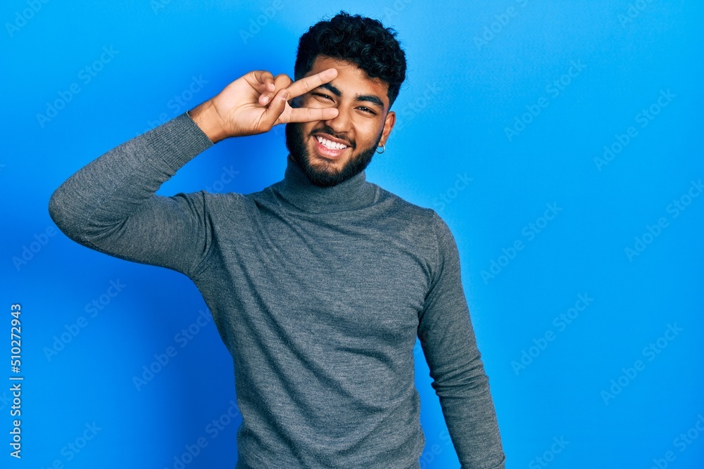 Arab man with beard wearing turtleneck sweater doing peace symbol with fingers over face, smiling cheerful showing victory Arab man with beard wearing turtleneck sweater doing peace symbol with fingers over face, smiling cheerful showing victory