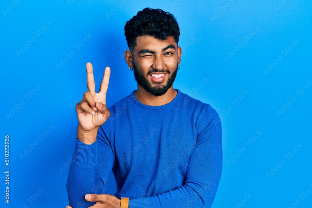 Arab man with beard wearing casual blue sweater smiling with happy face winking at the camera doing victory sign. number two. Arab man with beard wearing casual blue sweater smiling with happy face winking at the camera doing victory sign. number two.