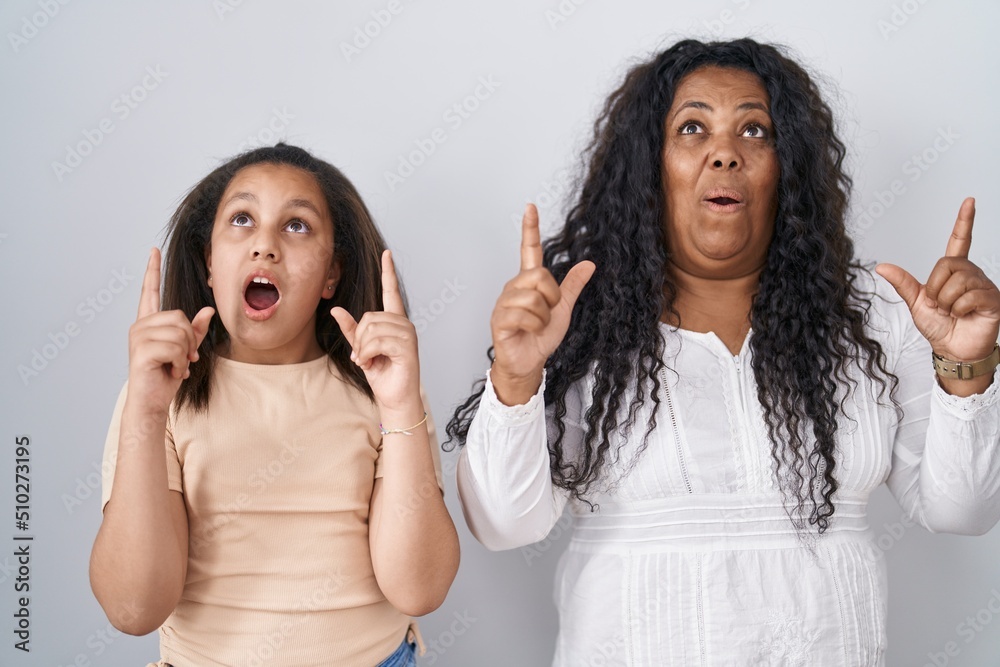Mother and young daughter standing over white background amazed and surprised looking up and pointing with fingers and raised arms. Mother and young daughter standing over white background amazed and surprised looking up and pointing with fingers and raised arms.