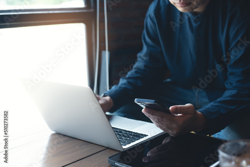 Young asian man sitting in coffee shop, using mobile phone browsing internet with laptop computer and digital tablet on table, male freelancer working at cafe