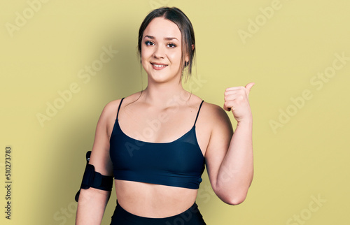 Young hispanic woman wearing sportswear smiling with happy face looking and pointing to the side with thumb up. © Krakenimages.com