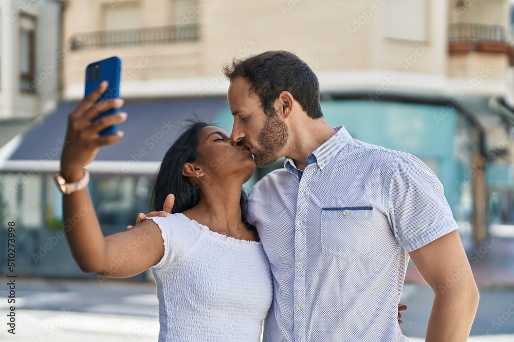 Man and woman interracial couple making selfie by smartphone kissing at street