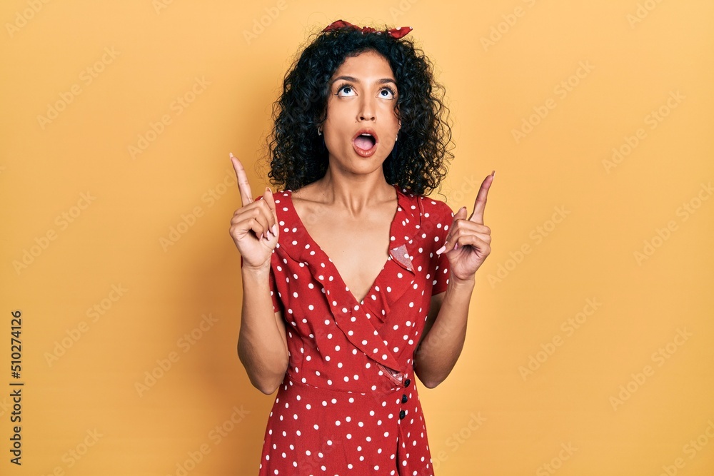 Young latin girl wearing summer dress amazed and surprised looking up and pointing with fingers and raised arms. Young latin girl wearing summer dress amazed and surprised looking up and pointing with fingers and raised arms.