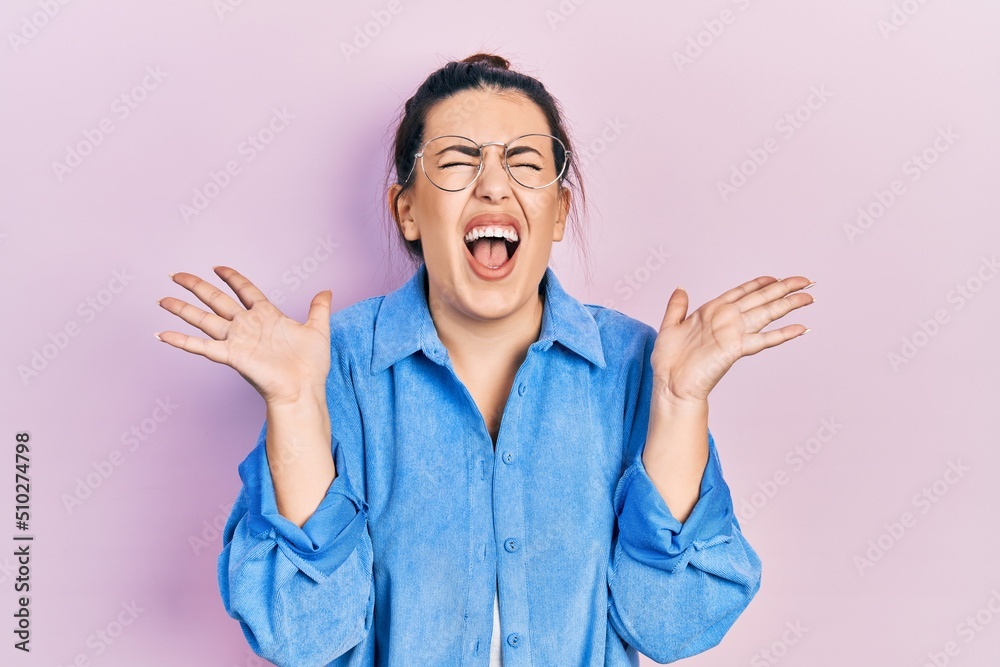 Young hispanic woman wearing casual clothes and glasses celebrating mad and crazy for success with arms raised and closed eyes screaming excited. winner concept Young hispanic woman wearing casual clothes and glasses celebrating mad and crazy for success with arms raised and closed eyes screaming excited. winner concept