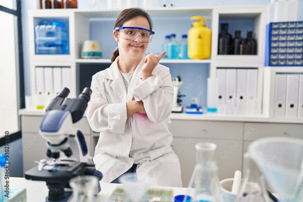 Hispanic girl with down syndrome working at scientist laboratory with a big smile on face, pointing with hand finger to the side looking at the camera. Hispanic girl with down syndrome working at scientist laboratory with a big smile on face, pointing with hand finger to the side looking at the camera.