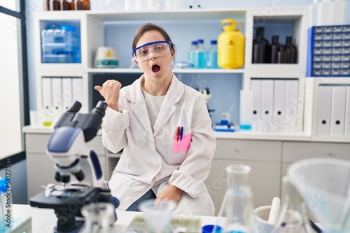 Hispanic girl with down syndrome working at scientist laboratory surprised pointing with hand finger to the side  open mouth amazed expression.