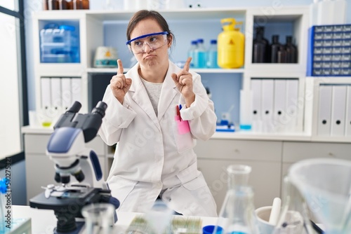 Hispanic girl with down syndrome working at scientist laboratory pointing up looking sad and upset  indicating direction with fingers  unhappy and depressed.