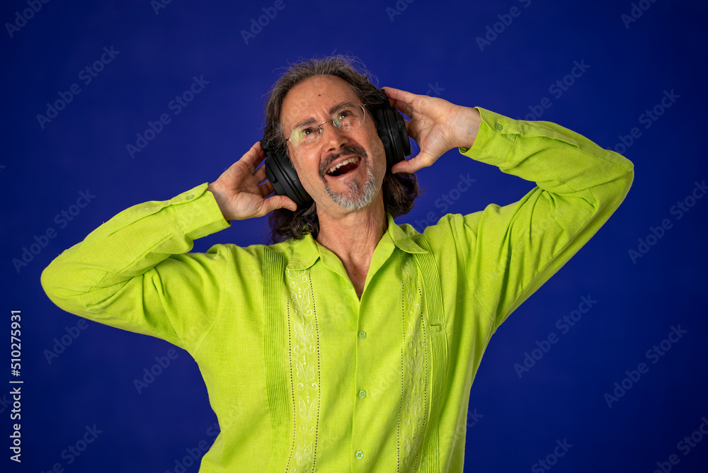 Happy mature bearded man 50s wear green shirt listen music in headphones dance fun enjoy relax isolated on solid blue background studio portrai Happy mature bearded man 50s wear green shirt listen music in headphones dance fun enjoy relax isolated on solid blue background studio portrai