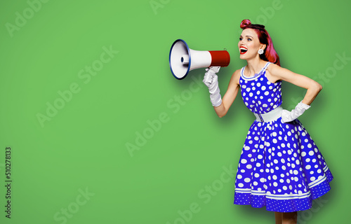 Purple haired woman holding red megaphone, shout advertising something. Girl in blue pin up style with mega phone loudspeaker. Green background with mock up. Female model in retro fashion dress.