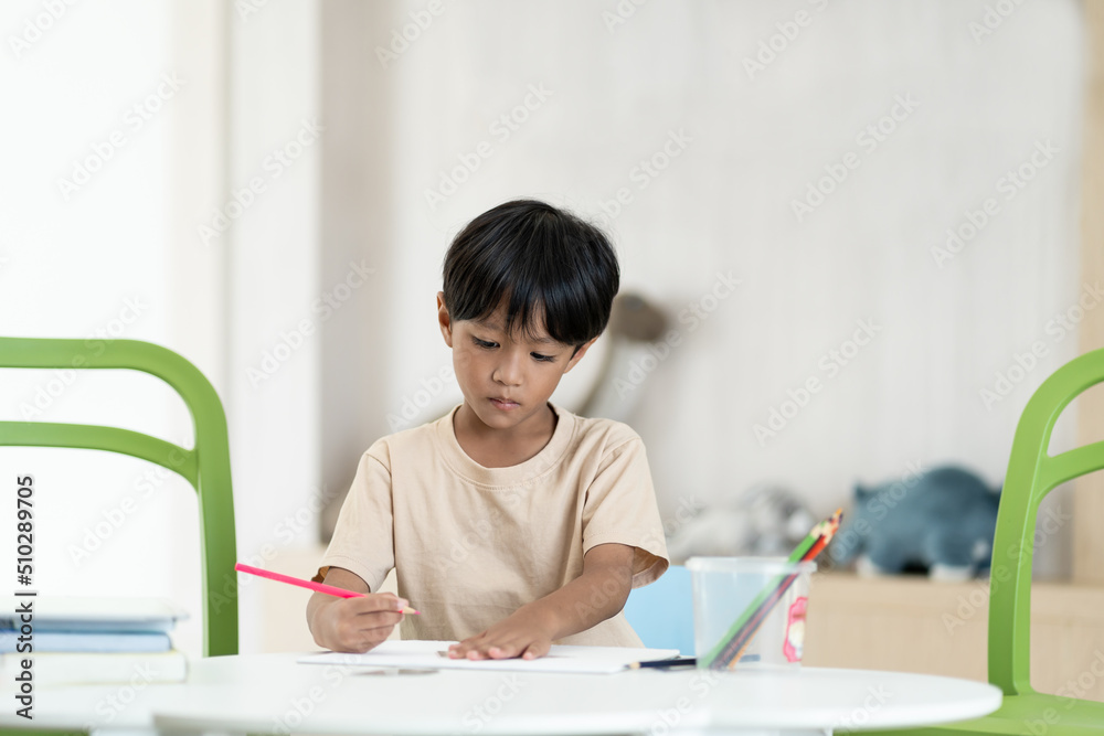Young asian child boy using pencil writing something in book at classroom. Asian son doing homework while sitting at desk at home. Kid learning by drawing. Education concept Young asian child boy using pencil writing something in book at classroom. Asian son doing homework while sitting at desk at home. Kid learning by drawing. Education concept