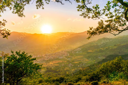 wonderful spring sunset in mountains with amazing view from highland among greeen tree branches to a valley with town, mountains and beautiful sunset on background