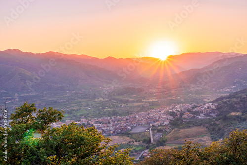 amazing highland landscape with scenic view from mountain with green branches and leaves on sides to a valley town with majectic mountains and scenic cloudy sunset on background