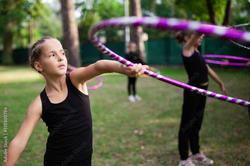 Girl doing exercise with hoop on rhythmic gymnastics training with other trainees outdoors in sports camp in summer