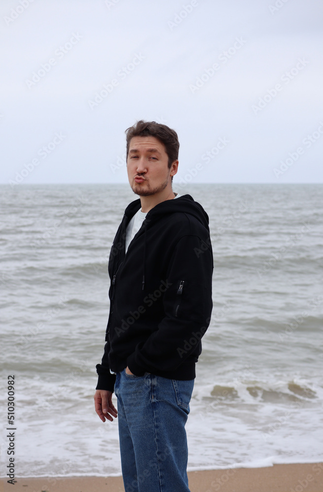 A young man in a black hoodie on the beach against the backdrop of the sea. Brown hair, blue eyes, smiling