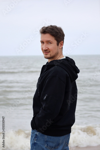 A young man in a black hoodie on the beach against the backdrop of the sea. Brown hair, blue eyes, smiling © Andrei Antipov