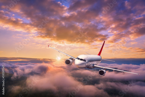 Airplane is flying above the clouds at sunset in summer. Landscape with passenger airplane, mountains, orange sky. Aircraft is taking off. Business travel. Commercial plane. Aerial view. Transport 