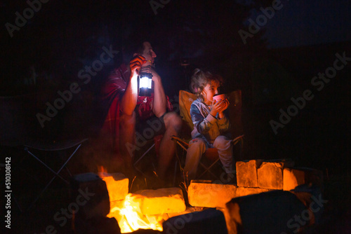 Dad and daughter sit at night by the fire in the open air in the summer in nature. Family camping trip, gatherings around the campfire. Father's Day, barbecue. Camping lantern and tent